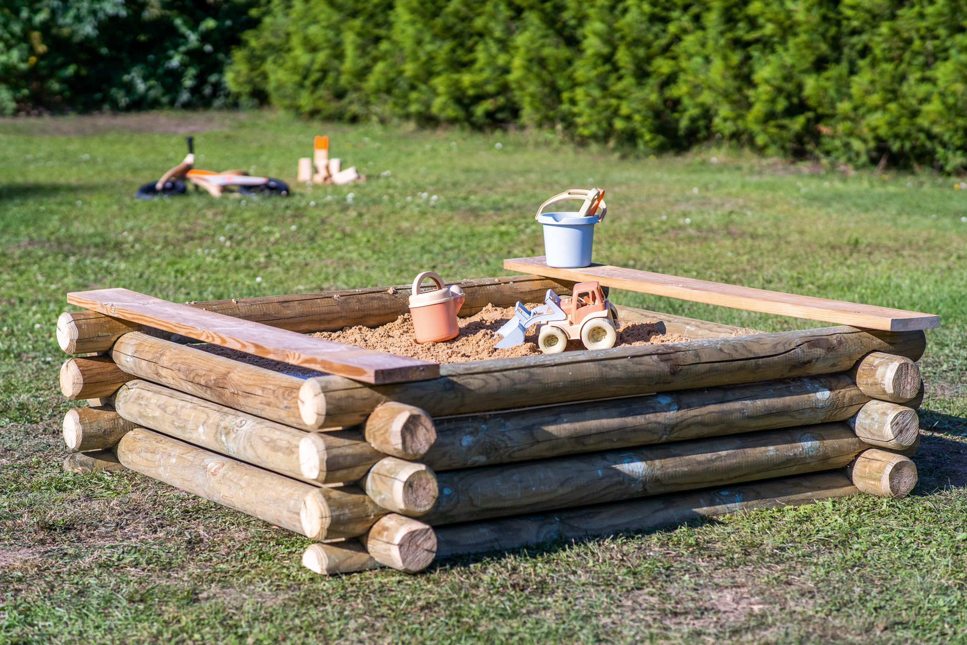 Bac à sable en bois avec bancs posé sur une pelouse, seaux et autres jouets.
